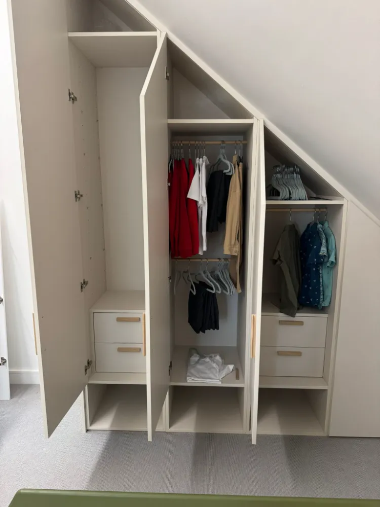 Fitted wardrobe with drawers built into sloped loft ceiling, doors open showing hanging clothes