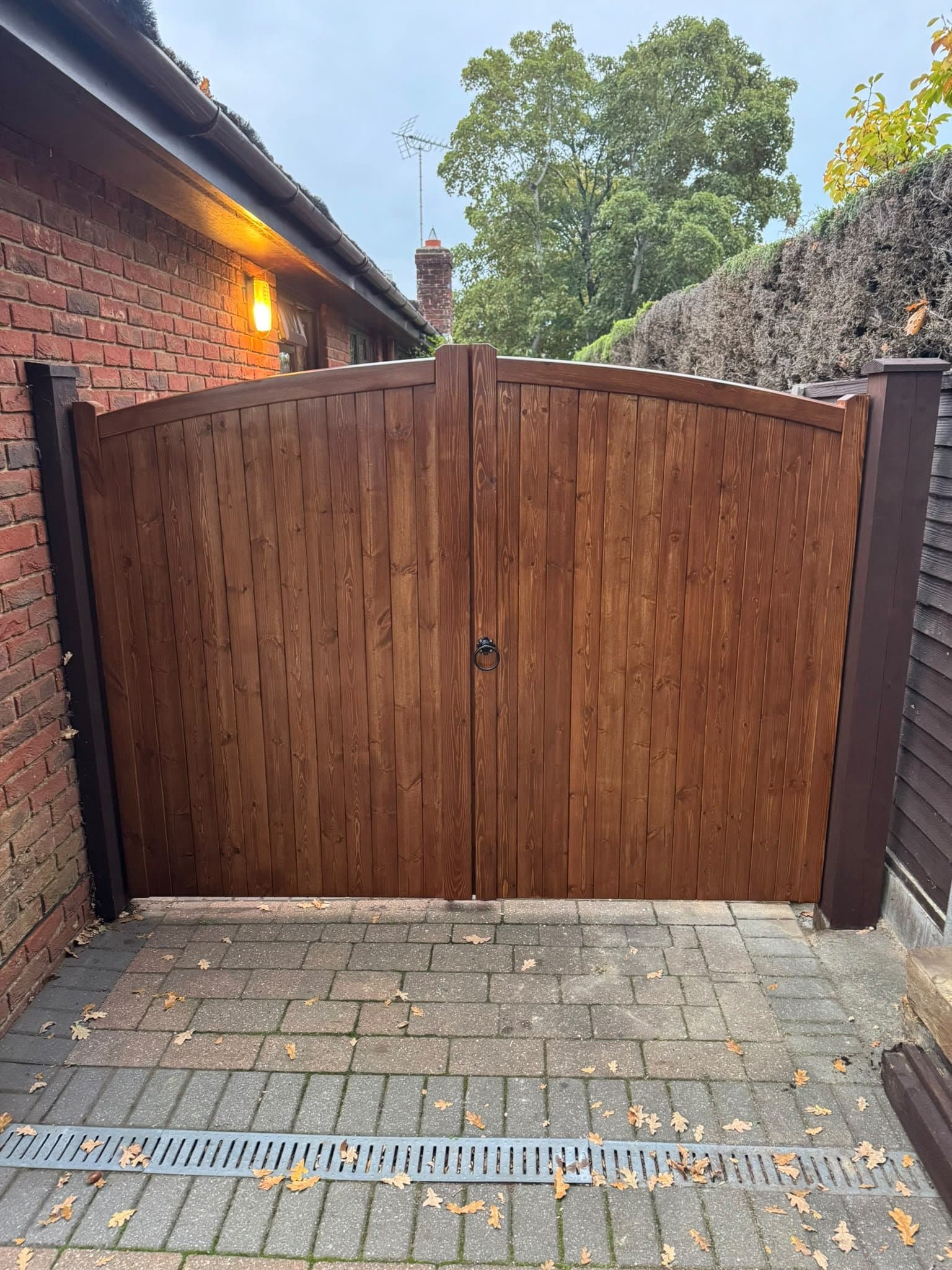Arched timber double driveway gates hung between stained posts beside a brick bungalow